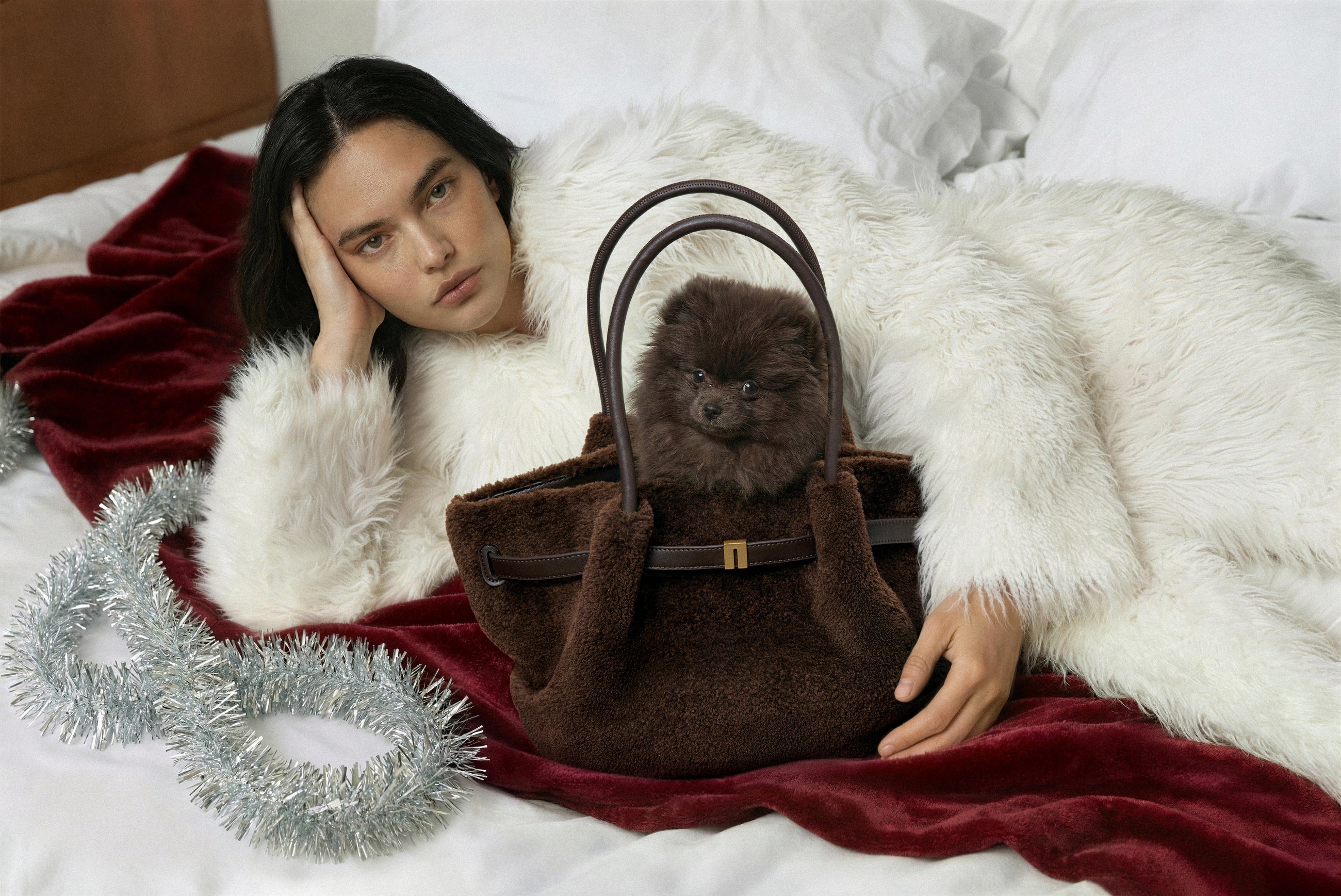 Person holding a brown handbag with a small dog inside, lying on a white blanket with red accents.